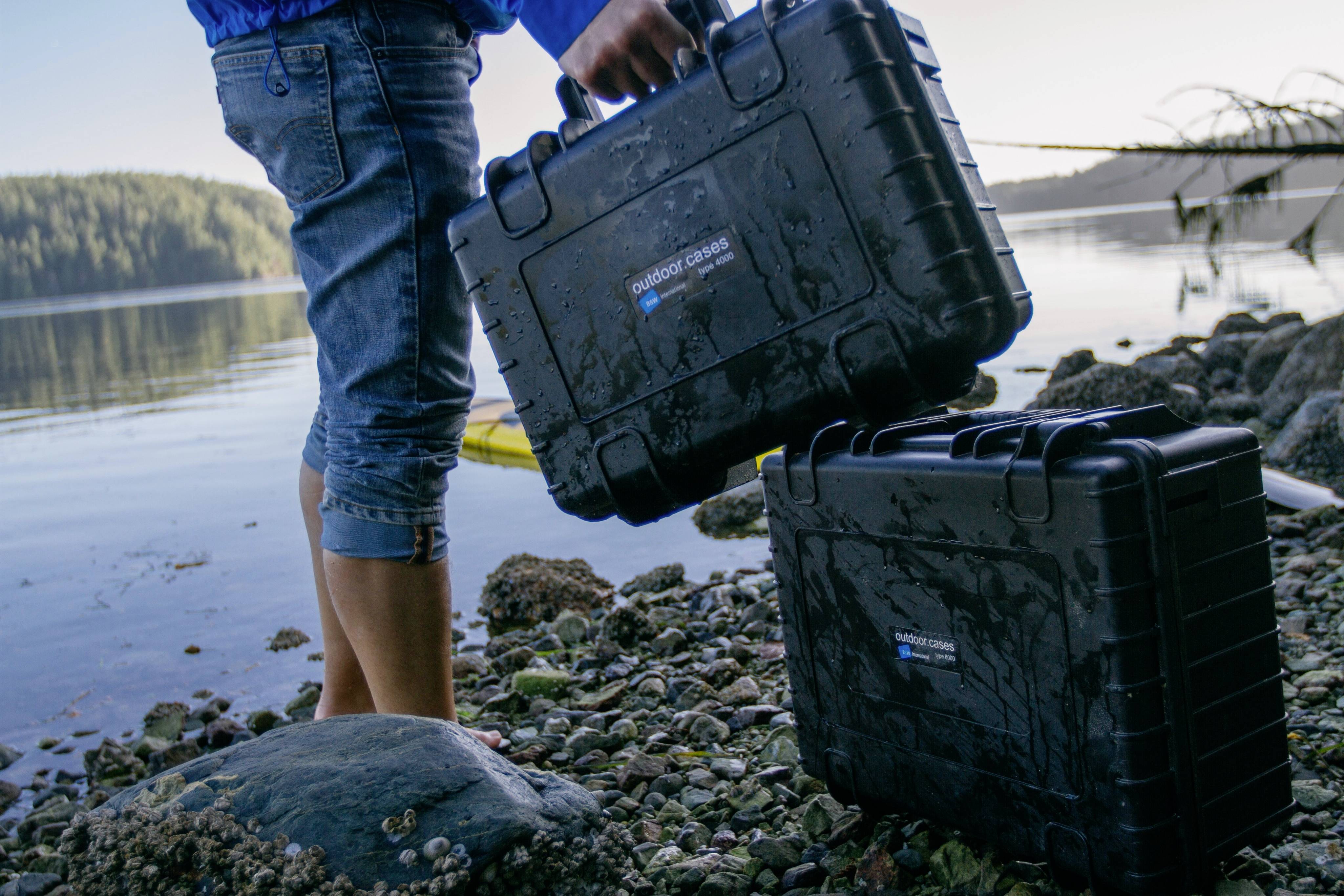 Une personne près de l'eau porte une valise noire imperméable. Une deuxième valise est posée sur le gravier. En arrière-plan : un lac et des arbres.