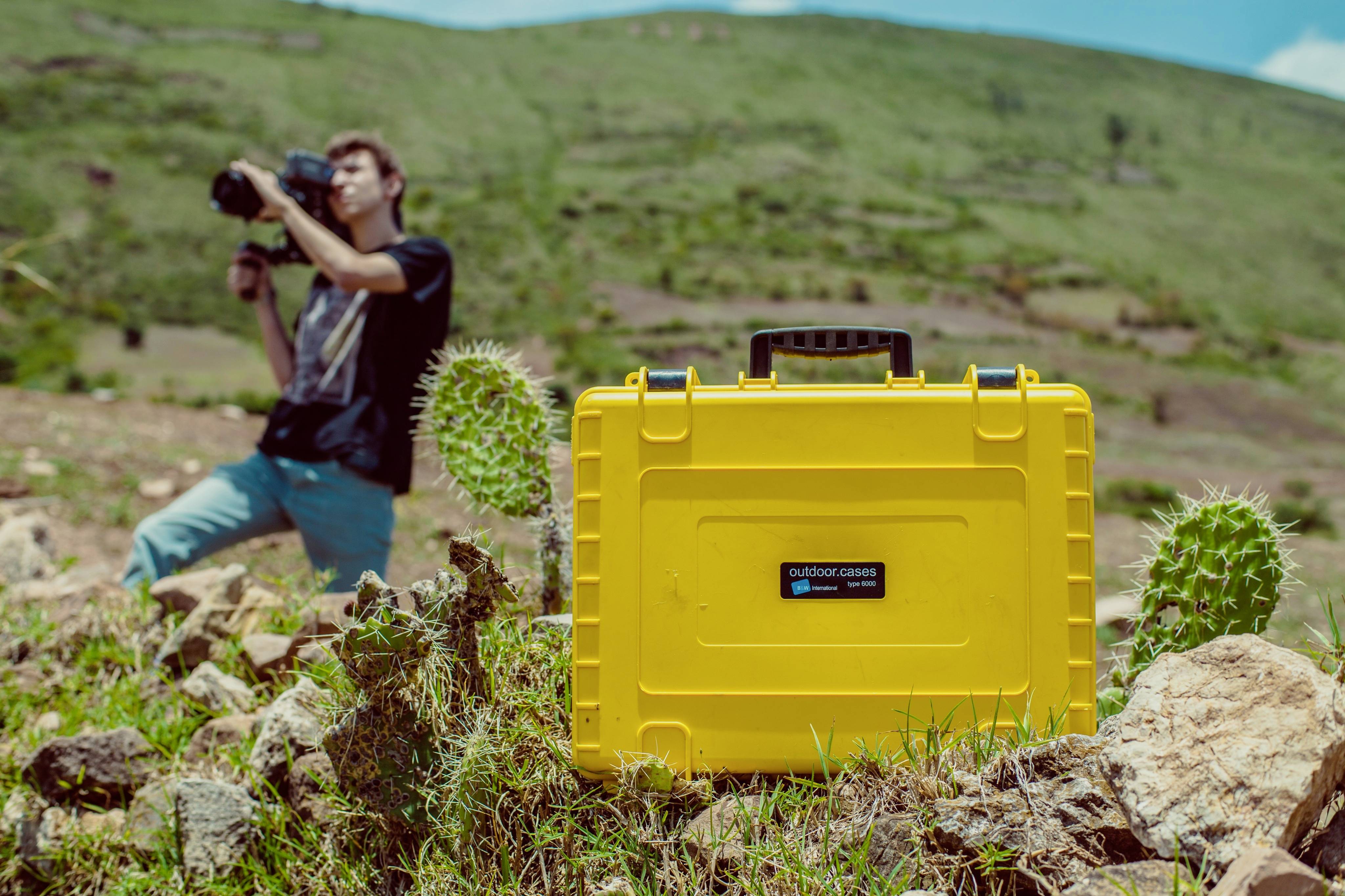 Valise jaune de plein air posée sur un sol rocailleux à côté de cactus ; en arrière-plan, une personne photographie le paysage.