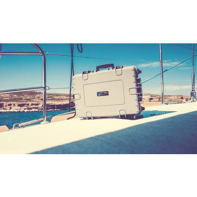 Une valise argentée est posée sur le pont d'un voilier naviguant sur une mer calme. La côte est visible en arrière-plan.