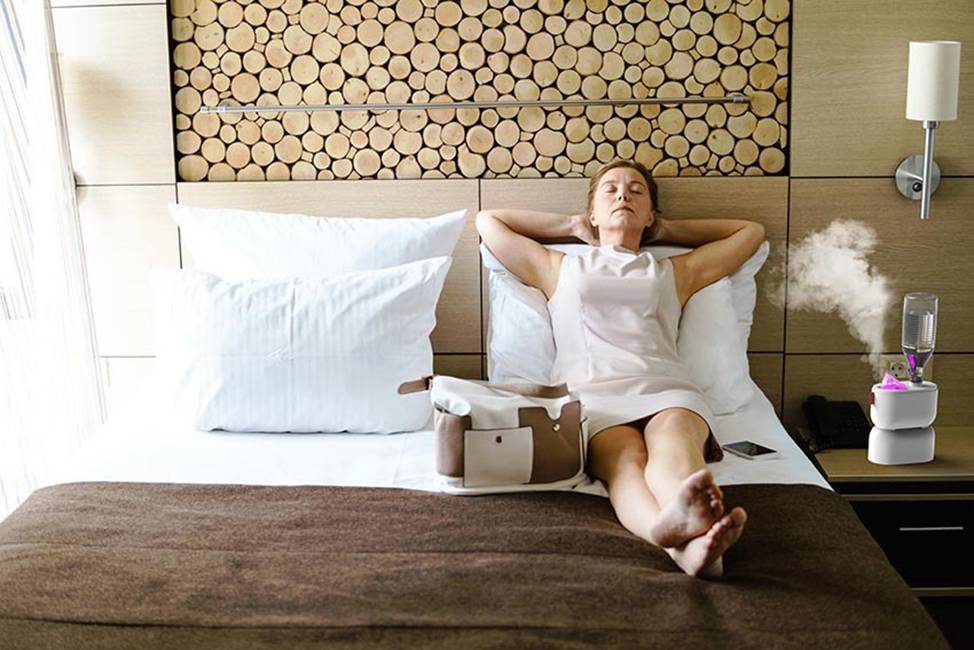 Une femme en robe blanche est allongée de façon détendue sur un lit d'hôtel. À côté d'elle, un sac et un humidificateur en marche.