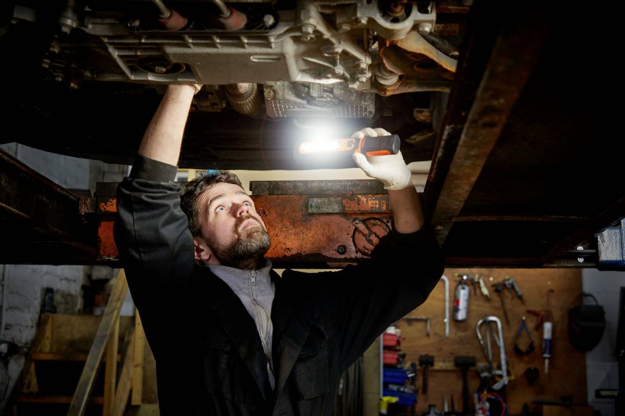 Un mécanicien automobile inspecte le soubassement d'un véhicule dans un atelier, tenant une lampe de travail pour une meilleure visibilité.