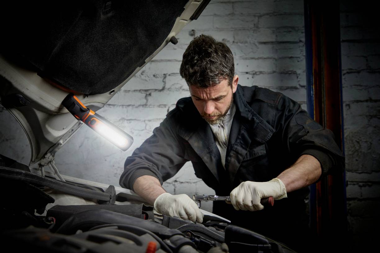 Un mécanicien répare le moteur d'une voiture avec des outils dans un atelier. Une lampe éclaire la zone de travail.