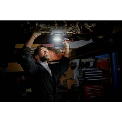 Un mécanicien travaille dans un atelier sous une voiture surélevée et éclaire la zone avec une lampe afin d'inspecter les pièces.
