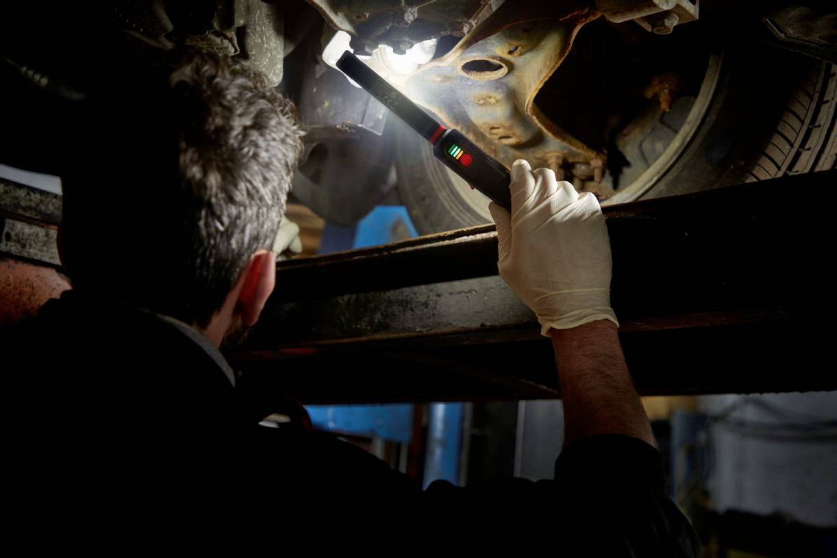 Un mécanicien portant des gants inspecte un véhicule sur un pont élévateur à l'aide d'une lampe torche. L'attention est portée sur le dessous du véhicule.