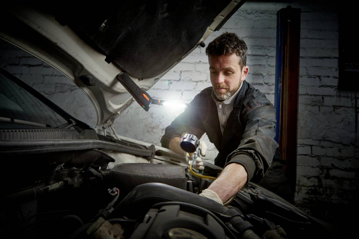 Un mécanicien travaille sous le capot d'une voiture, éclairé par une lampe de travail. Il porte une tenue de travail et des gants.