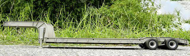 Un remorque grise à deux essieux est stationnée sur une route, entourée de buissons verts et d'herbe en arrière-plan.