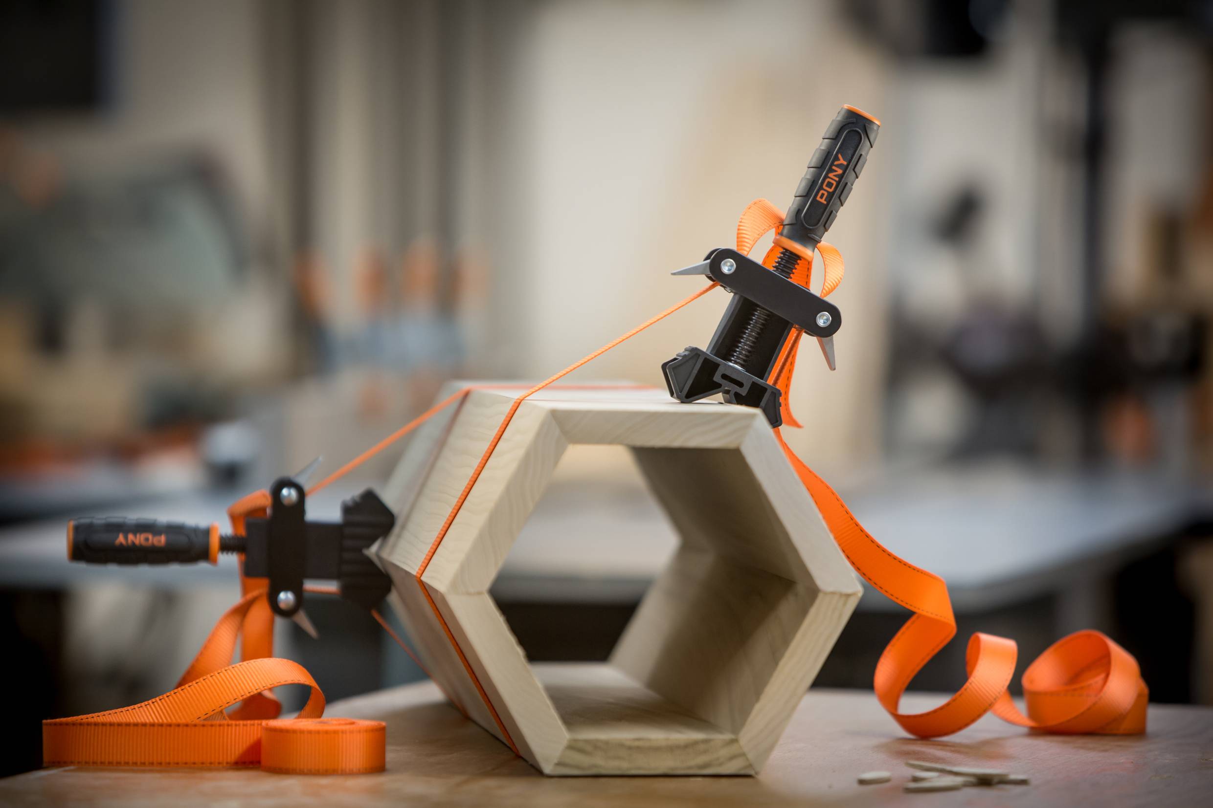 Un cube en bois de forme hexagonale, fixé avec une sangle orange, est posé sur l'établi. À l'arrière-plan, un environnement d'atelier flou.