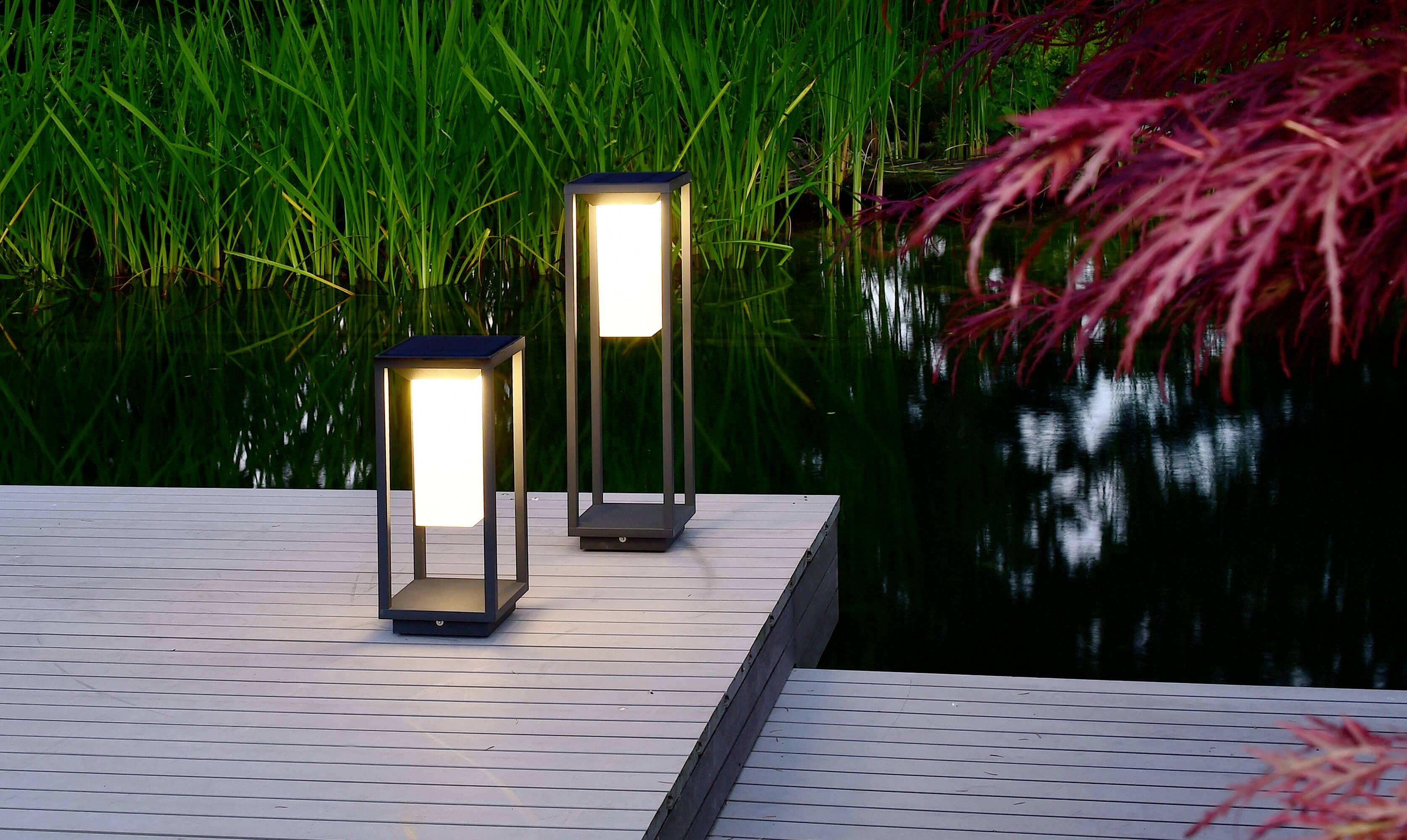 Deux lampadaires modernes sur une terrasse en bois près de l'eau, entourés de plantes vertes et de feuilles rouges en arrière-plan.