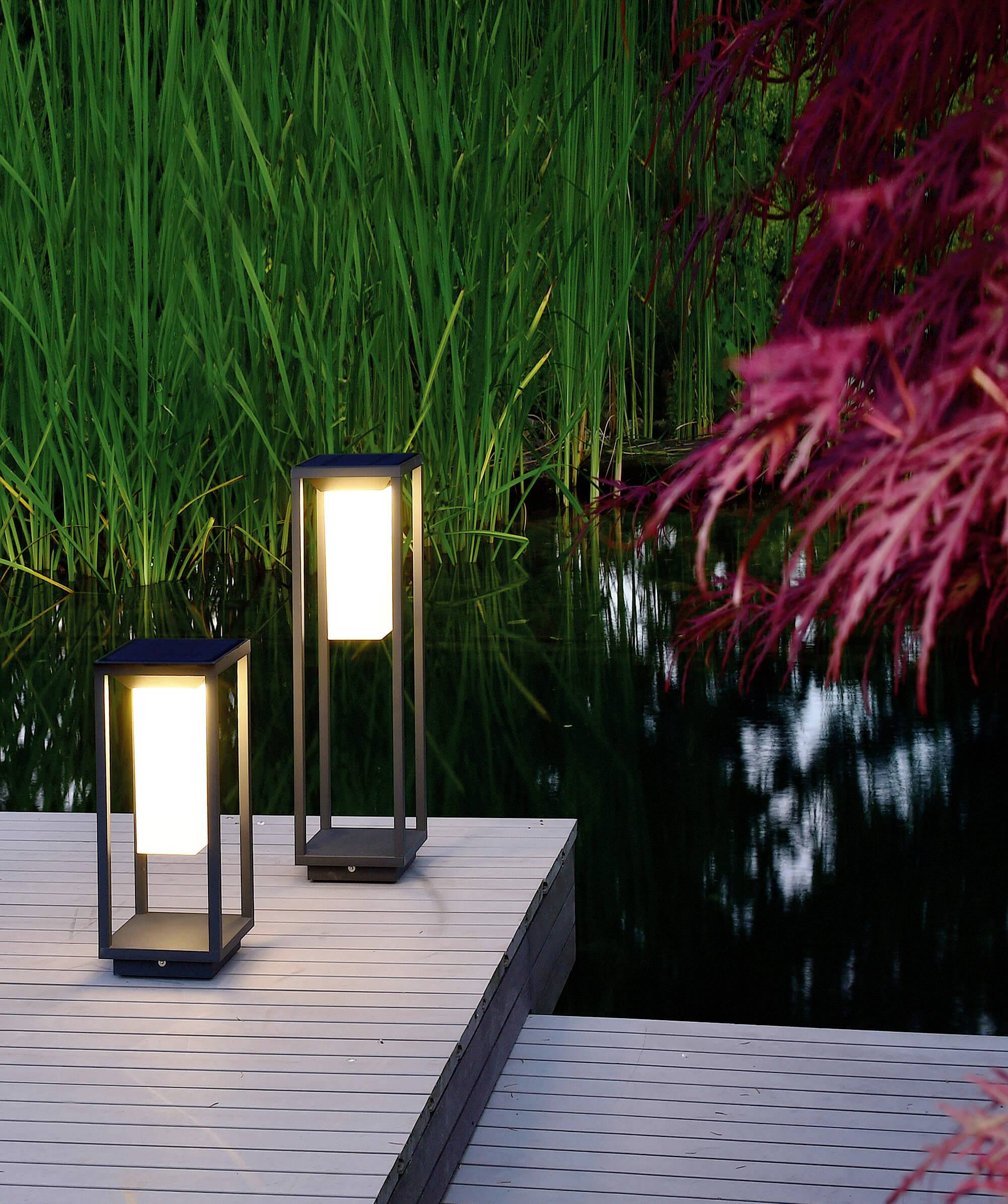 Deux lanternes lumineuses sur une passerelle en bois à côté d'un étang, entourées d'herbe haute verte et de feuilles rouges en arrière-plan.