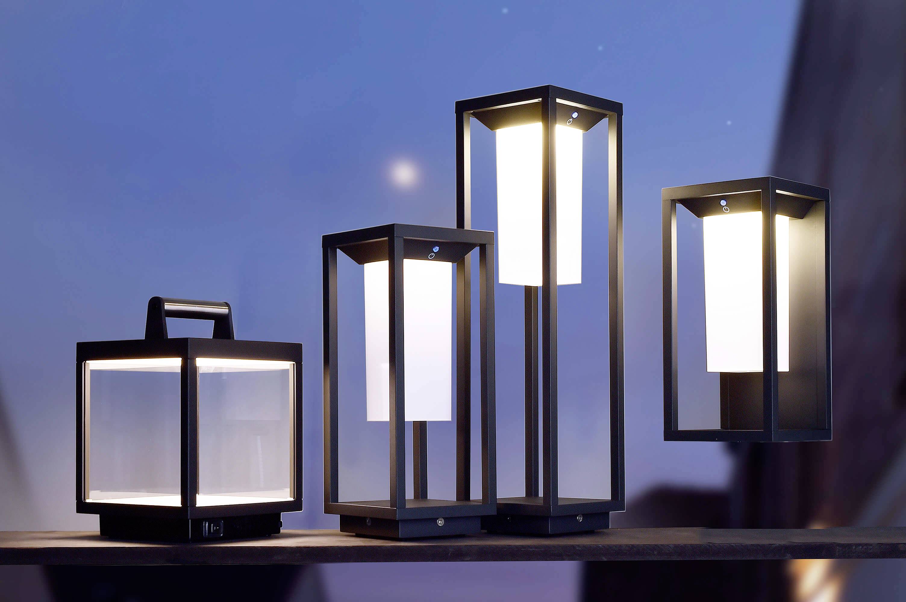 Quatre lanternes modernes, carrées, en métal noir sont disposées sur une surface en bois au crépuscule, toutes équipées de lumières LED chaudes et lumineuses.