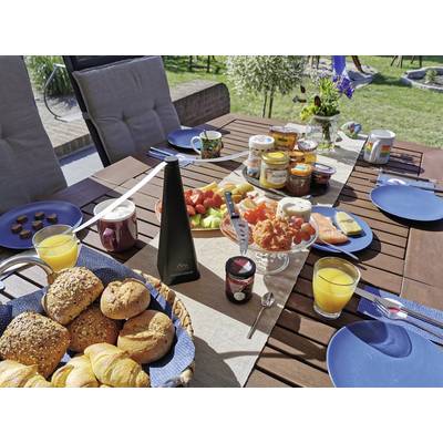 Table du petit-déjeuner en plein air avec petits pains, croissants, charcuterie, fruits, confiture et jus d'orange, entourée d'un décor de jardin verdoyant.