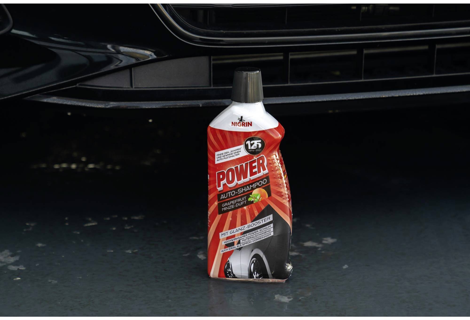 Une bouteille de shampoing pour voiture rouge est posée sur un sol humide devant un pare-chocs. La bouteille a un bouchon noir.