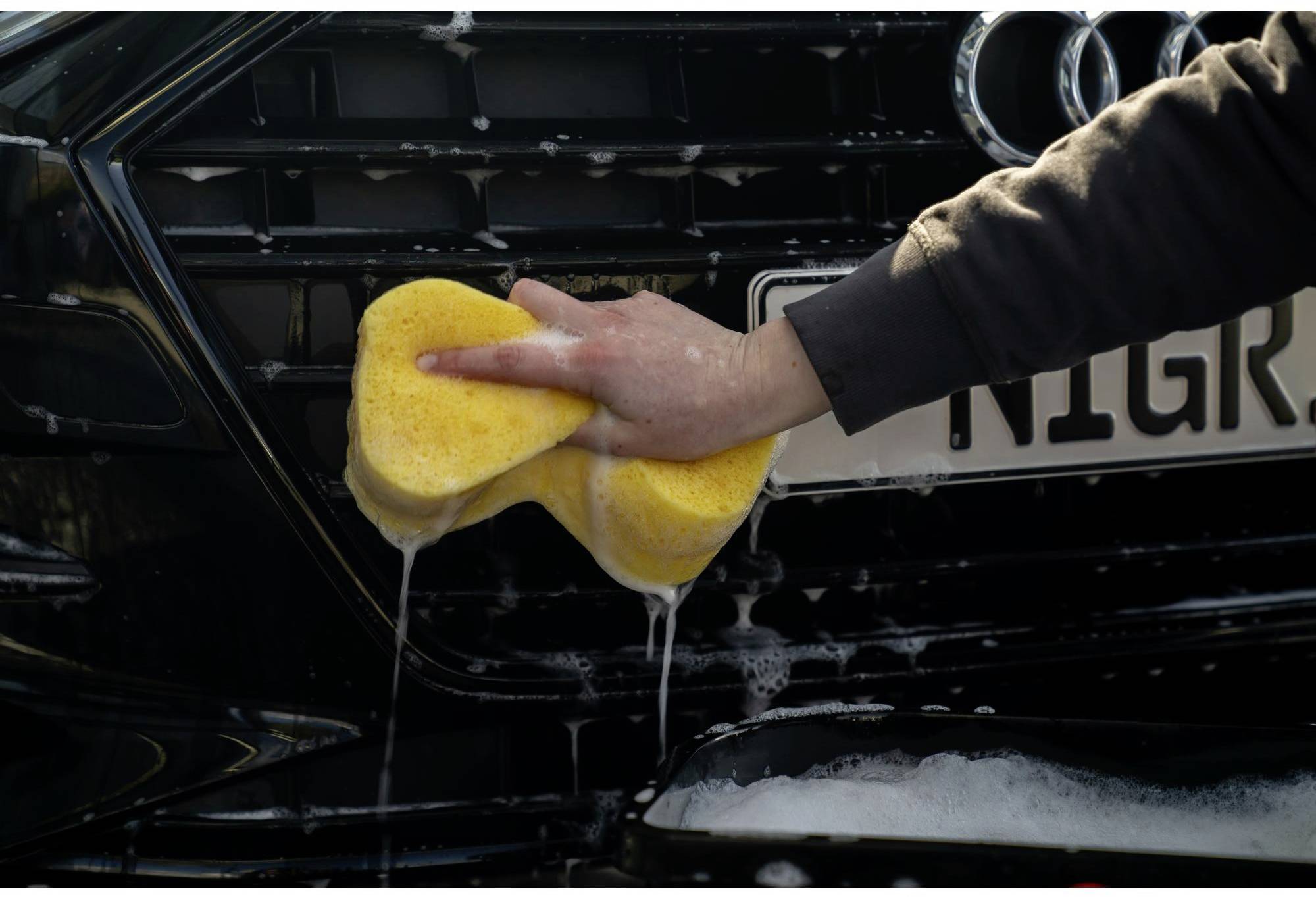Une personne lave la calandre d'une voiture avec une éponge jaune. De la mousse et de l'eau sont visibles, la plaque d'immatriculation est partiellement cachée.