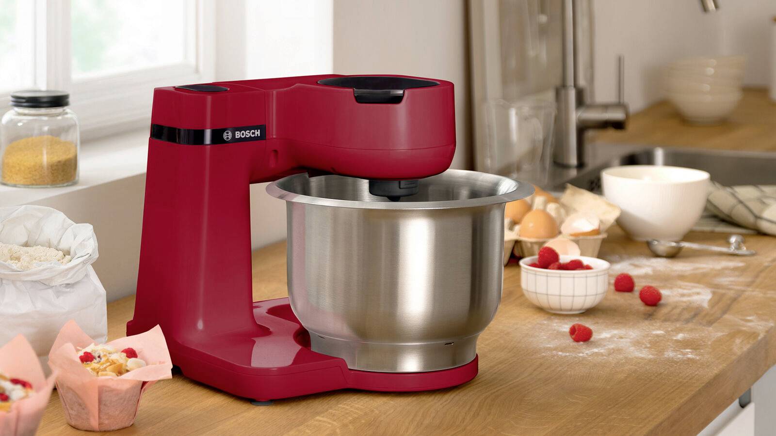 Machine à mixer rouge avec un bol en acier inoxydable sur un plan de travail en bois. À côté de la machine se trouvent de la farine, des œufs et des framboises.