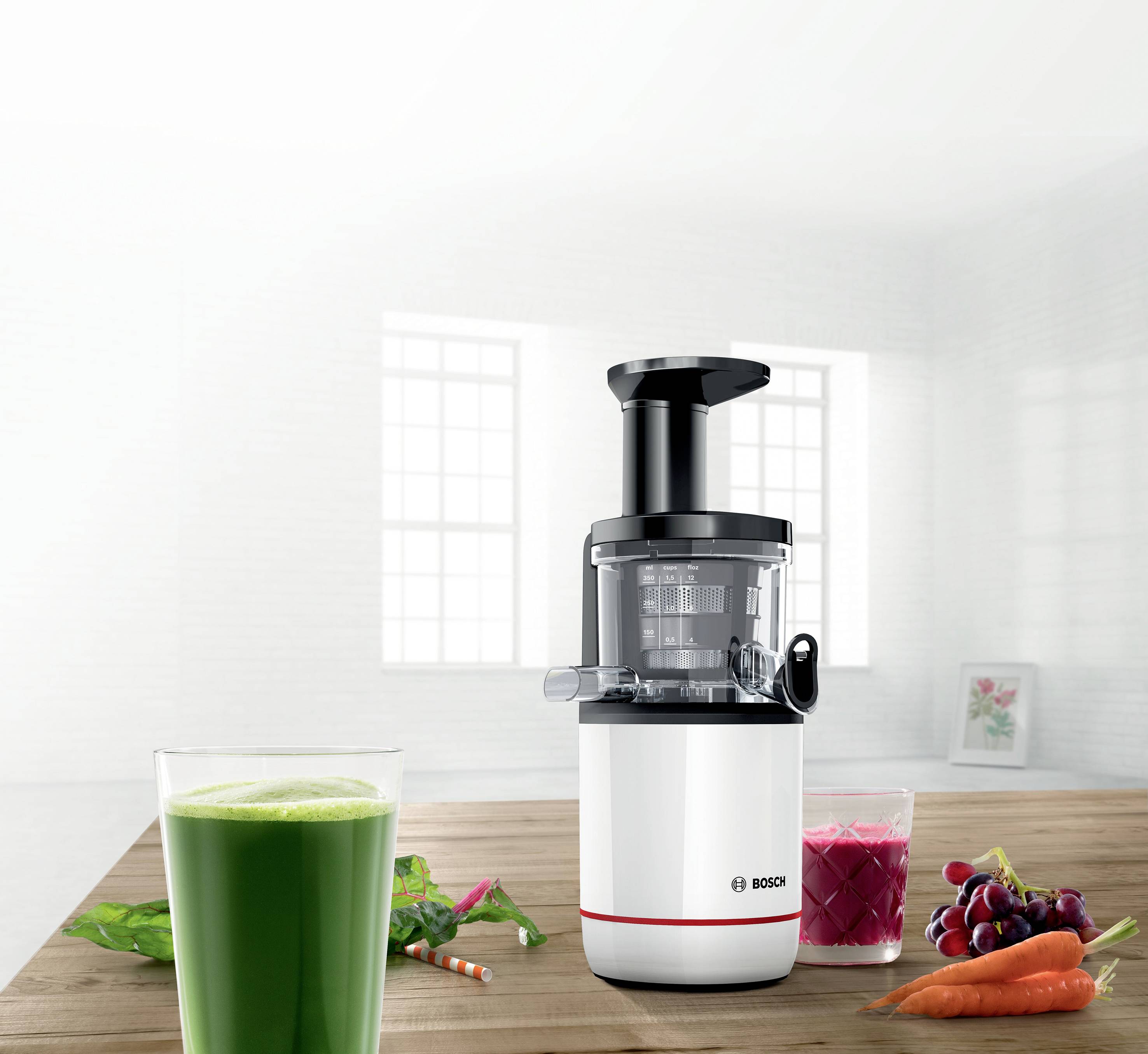 Extracteur de jus sur une table en bois avec deux verres de jus, l'un vert et l'autre rouge. À l'arrière-plan, une cuisine lumineuse avec de grandes fenêtres.