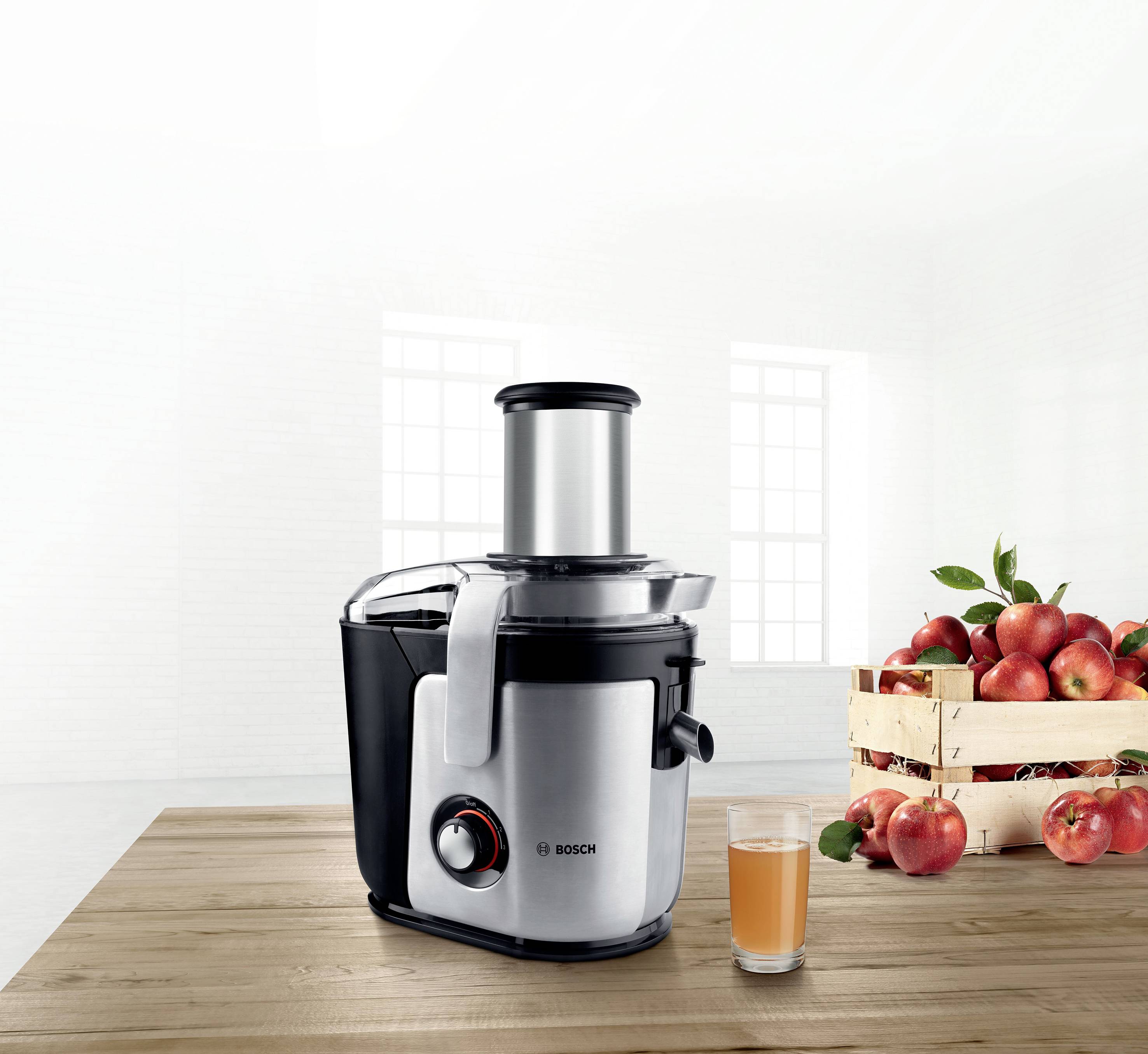 Un extracteur de jus posé sur une table en bois, à côté un verre de jus fraîchement pressé et, en arrière-plan, un panier rempli de pommes rouges.