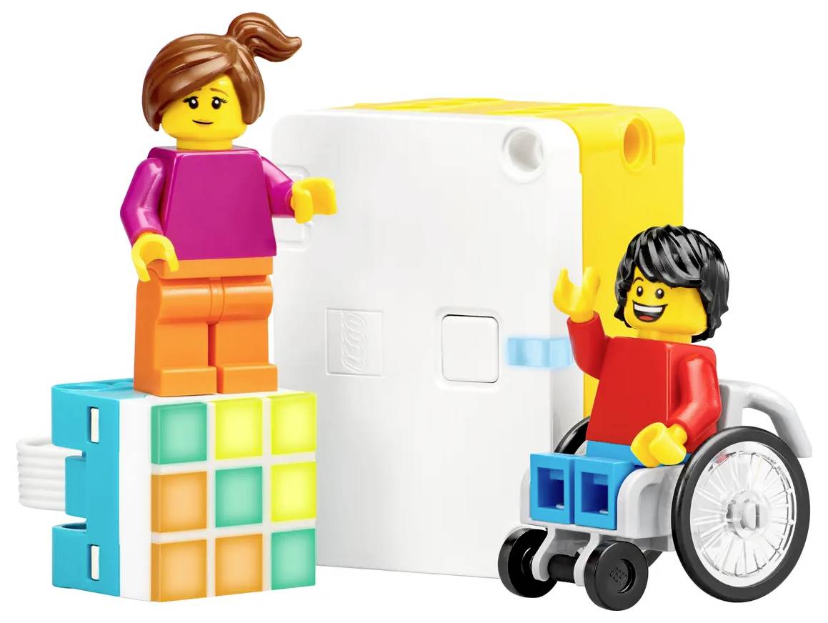 Une figure se tient sur un cube coloré. Une autre est assise dans un fauteuil roulant, toutes deux sourient. Des blocs blancs sont visibles en arrière-plan.