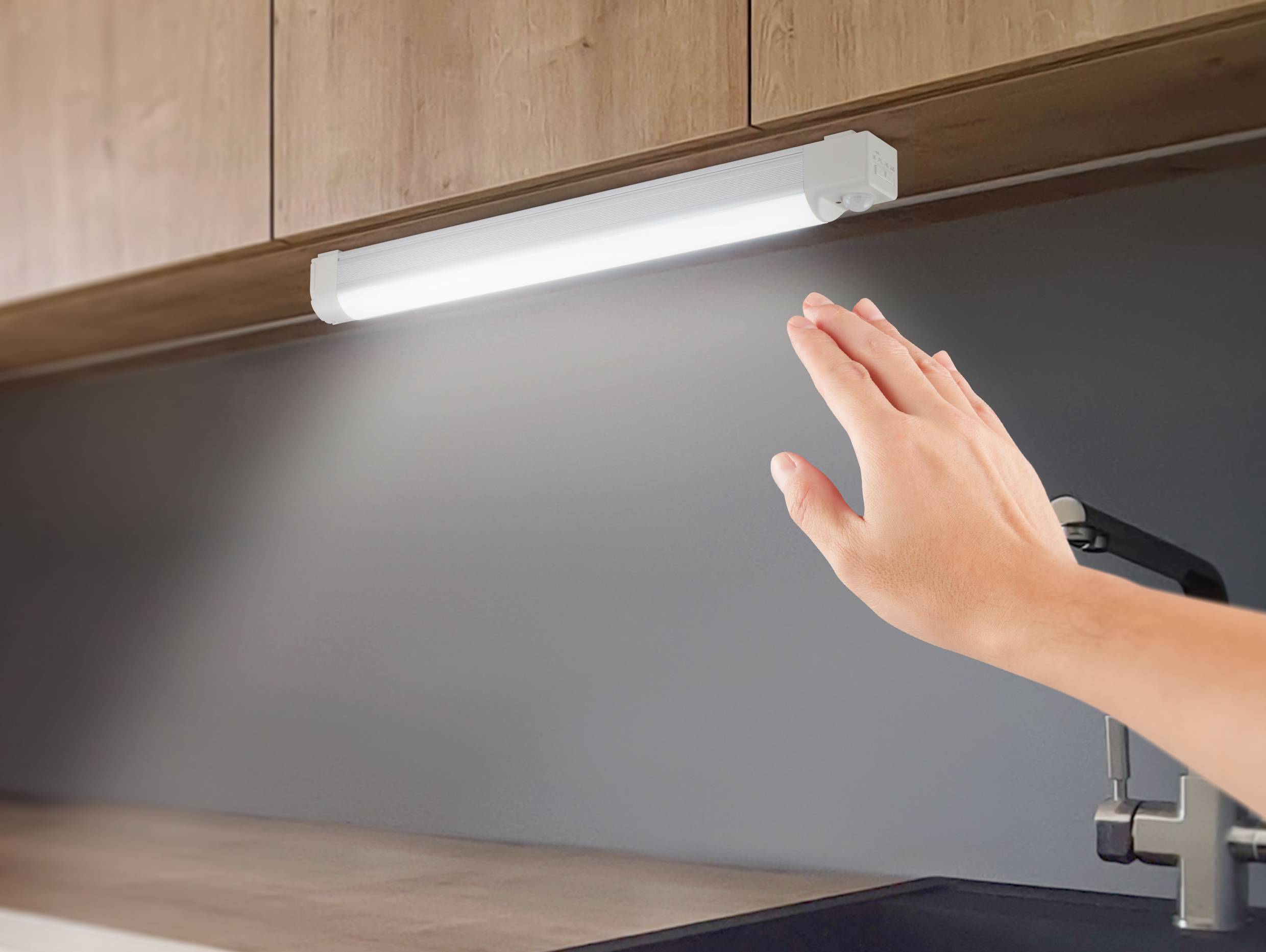 Une main survole une lampe LED allumée sous un placard de cuisine en bois, près d'un robinet.