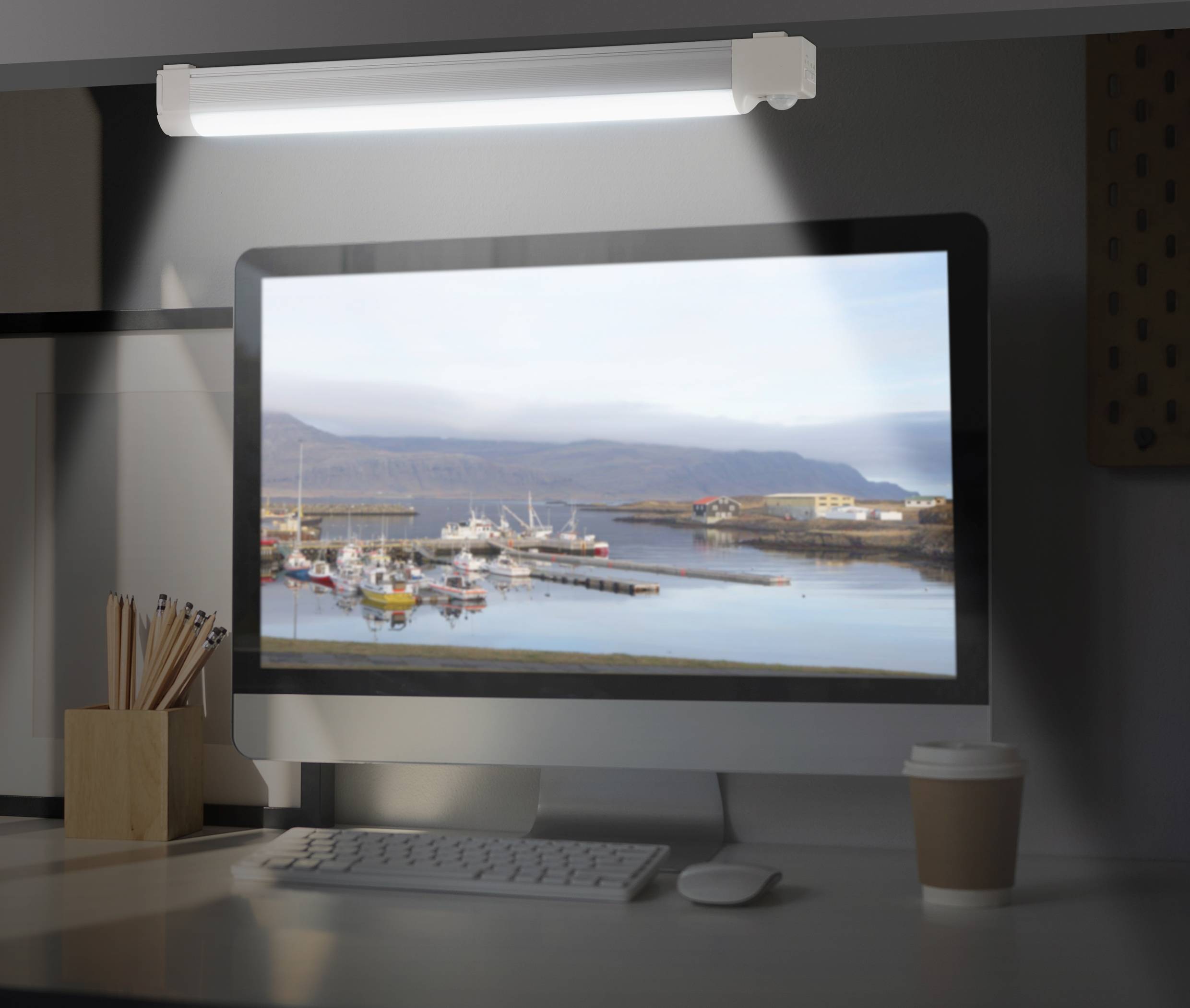 Un bureau avec un écran d'ordinateur affichant une image d'un port avec des bateaux et des montagnes en arrière-plan. Sur la table, il y a des stylos et une tasse de café.