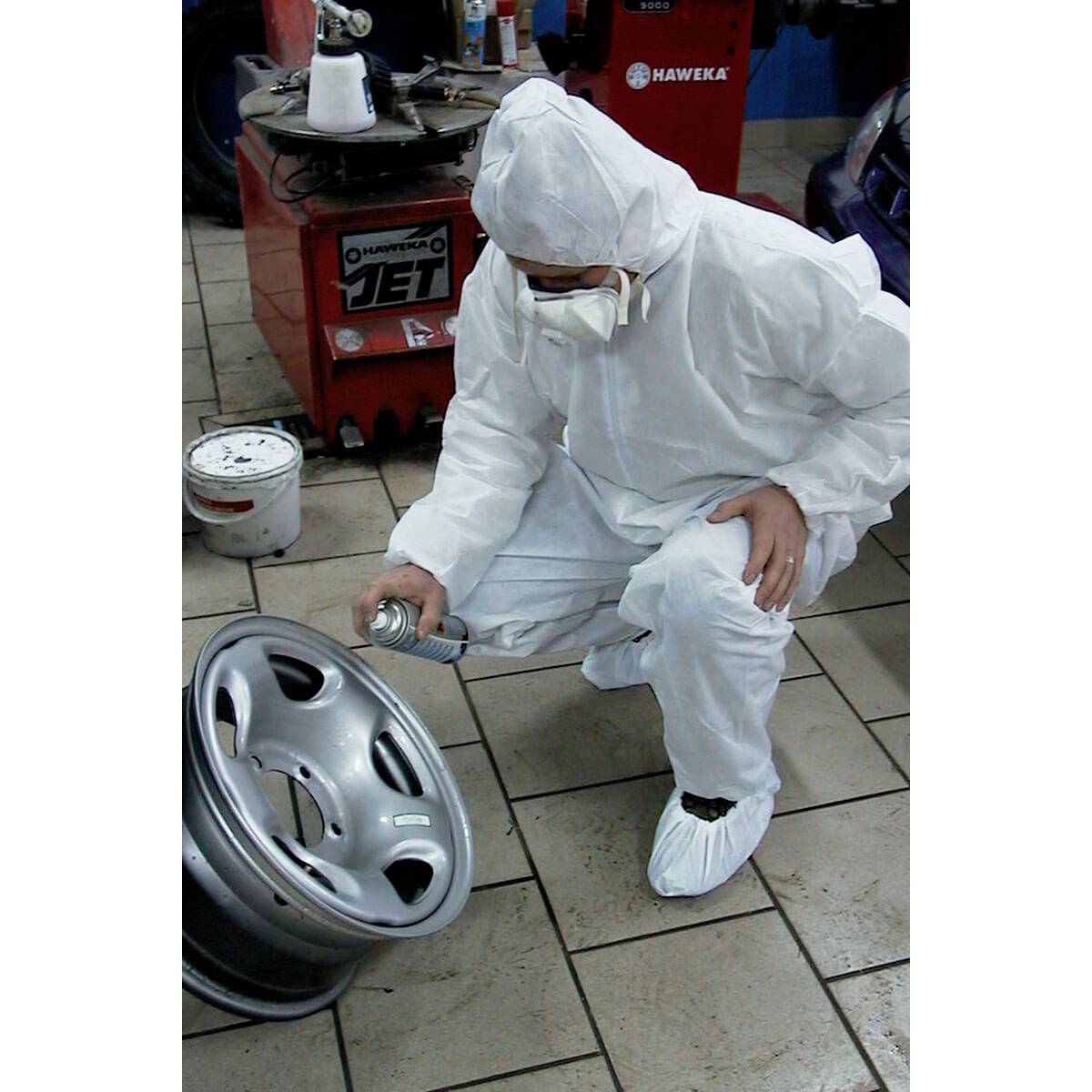 Une personne en combinaison de protection pulvérise de la peinture argentée sur une jante de voiture dans un atelier, afin de la peindre.