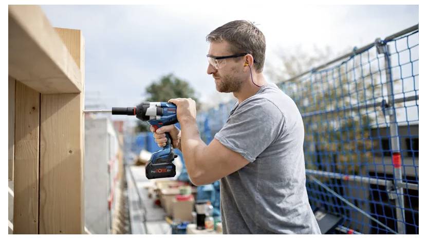 Un homme portant des lunettes de sécurité utilise une perceuse électrique sur une poutre en bois sur un chantier de construction, avec des barrières de sécurité et de l'équipement tout autour.