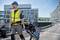 Un ouvrier porte un gilet de sécurité et un casque. Il tire un sac à outils sur un chariot à roulettes le long d'un trottoir devant des immeubles de bureaux modernes.