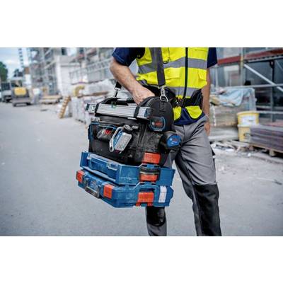 Travailleur portant un gilet de sécurité, transportant une boîte à outils sur un chantier. L'arrière-plan montre des échafaudages et des piles de matériaux de construction.
