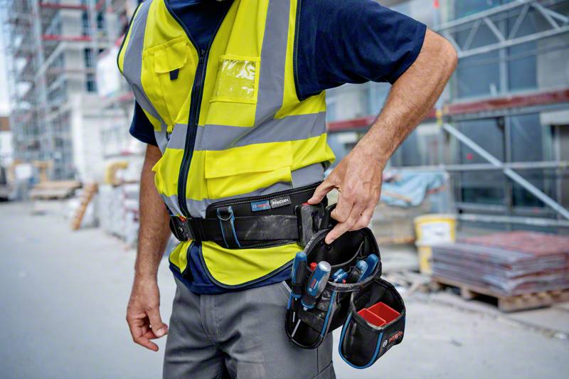 Un ouvrier sur un chantier porte un gilet de sécurité jaune et une ceinture à outils, tandis qu'il en sort des outils.