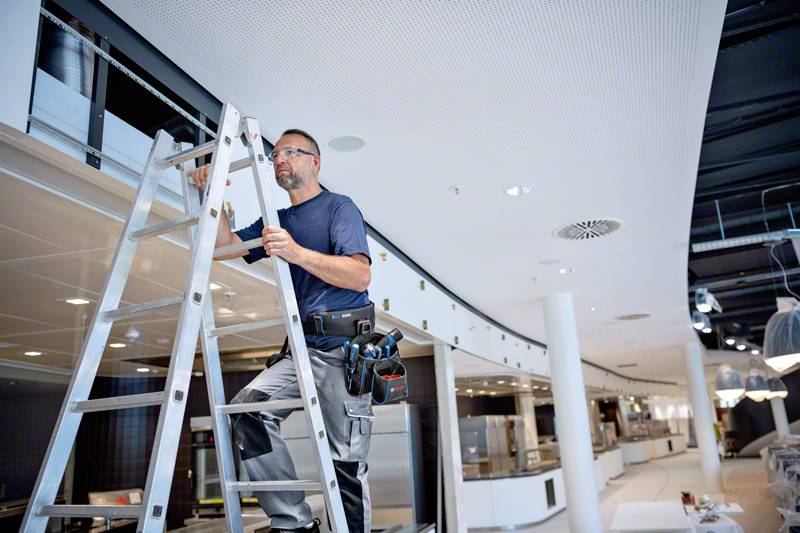 Un artisan sur une échelle répare un plafond dans un bâtiment moderne et lumineux, équipé d'outils à sa ceinture.