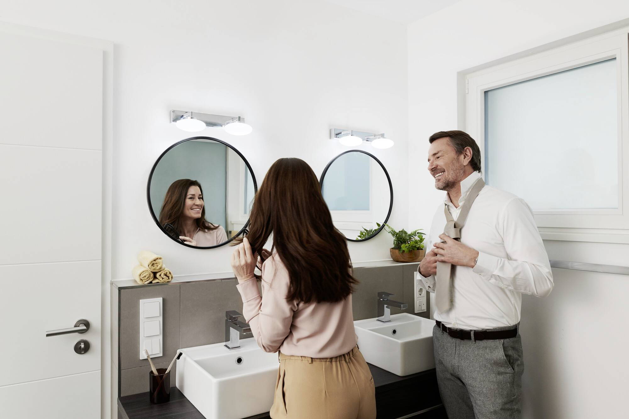 Une femme et un homme sont côte à côte dans une salle de bain, chacun devant un miroir. La femme arrange ses cheveux, l'homme noue sa cravate.