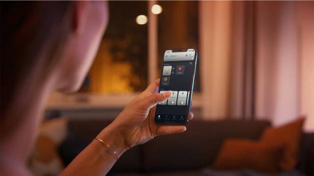 Une personne utilise une application mobile pour contrôler des fonctions domotiques, avec en arrière-plan un salon éclairé de manière tamisée.