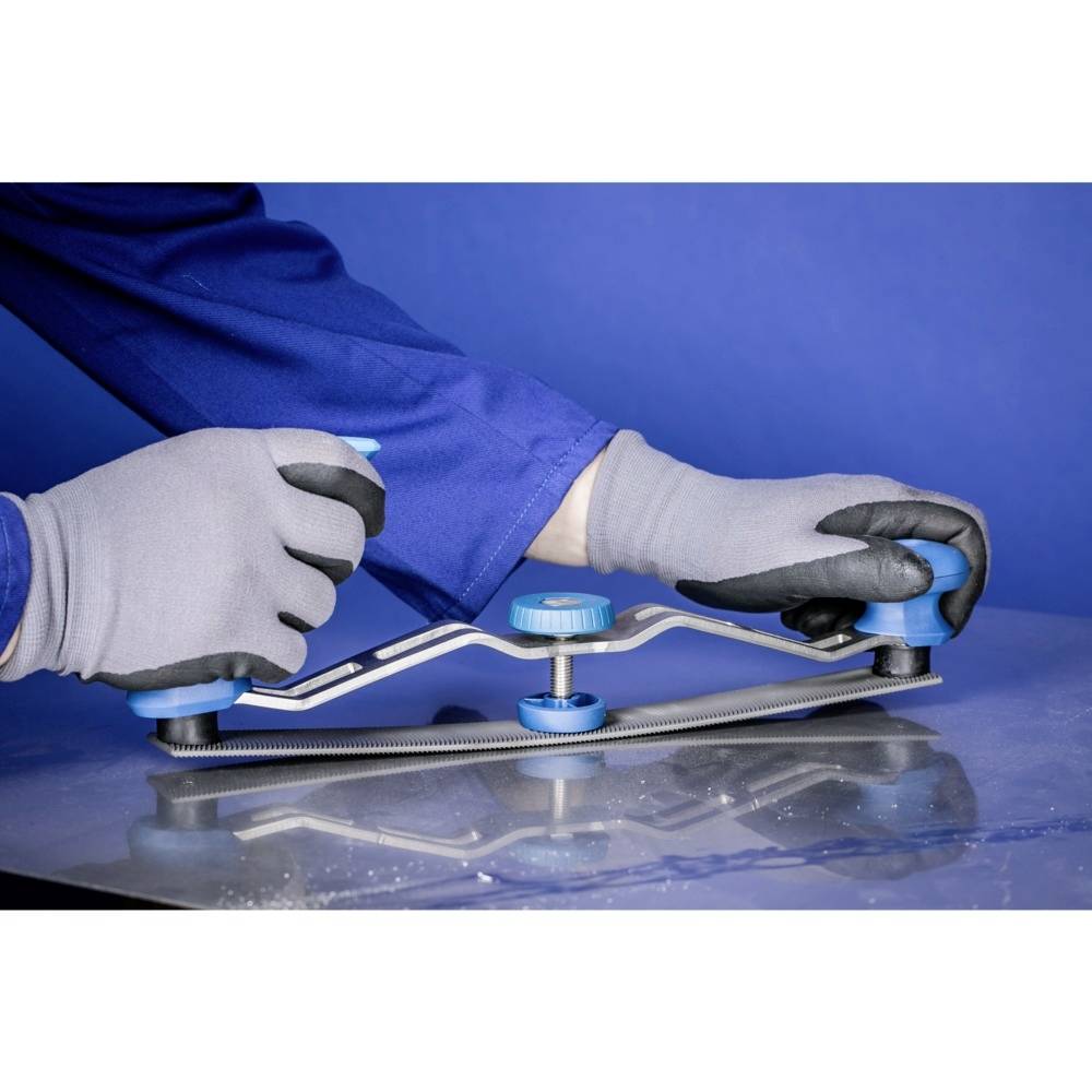 Une personne vêtue de vêtements de travail bleus et portant des gants gris serre une tôle métallique avec un outil bleu sur une table.