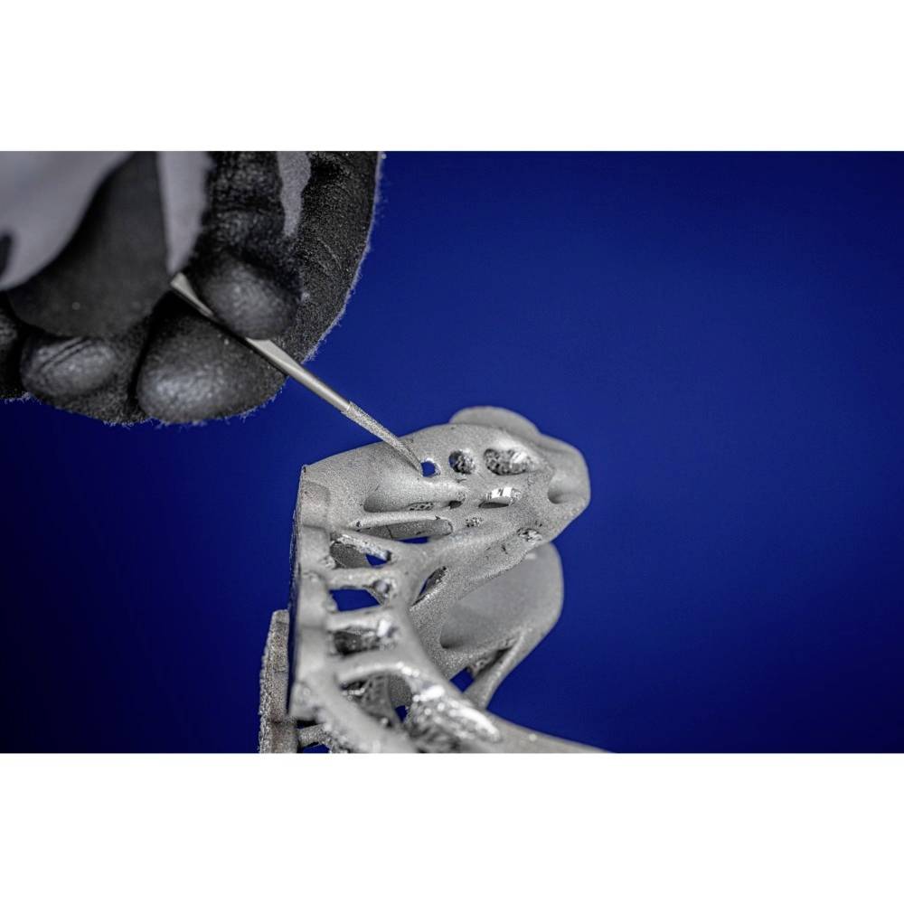 Une main gantée de noir manipule un élément métallique complexe imprimé en 3D à l'aide d'un outil, sur un fond bleu.