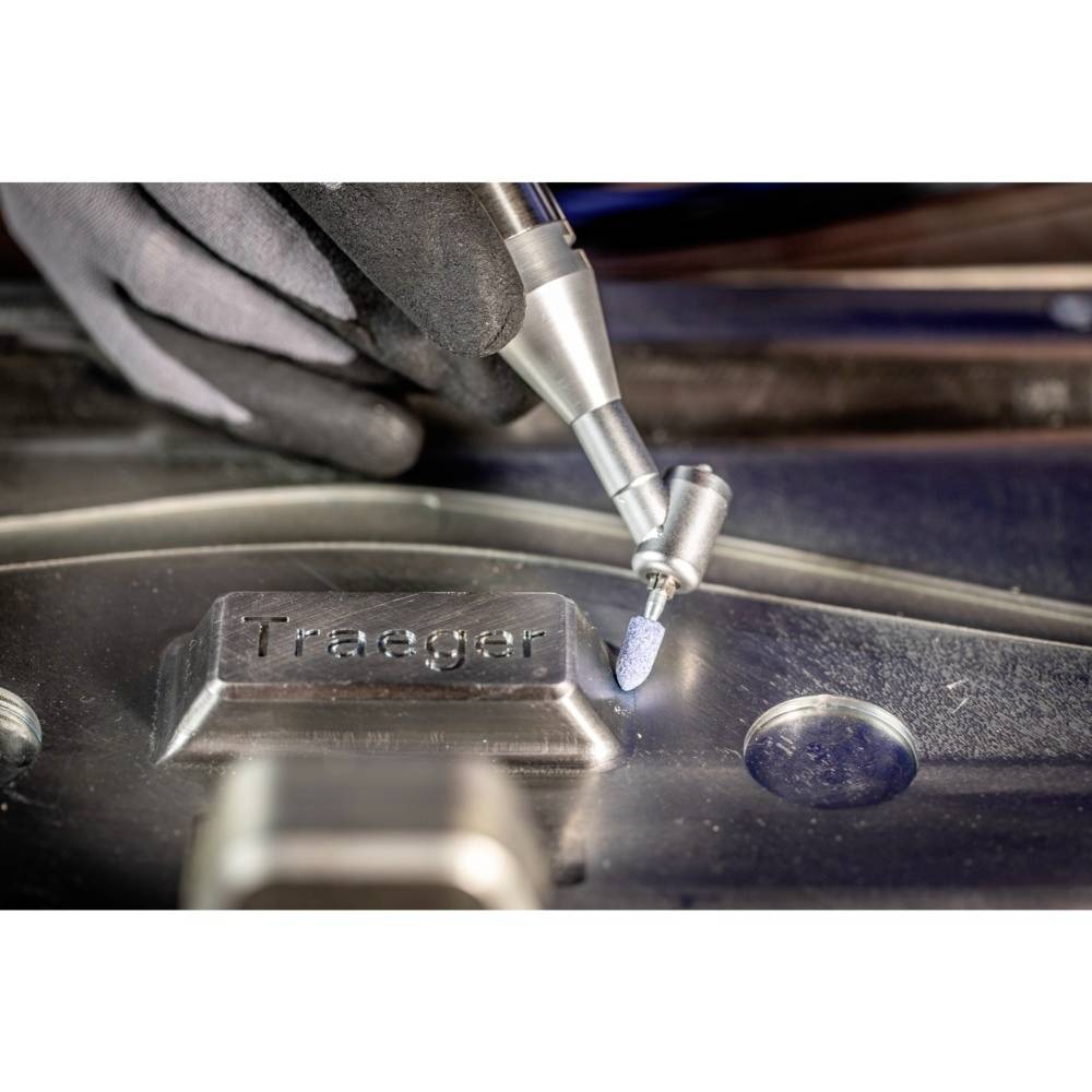 Un artisan traite une surface métallique avec un outil de meulage, sur laquelle est gravé 'Traeger'. Des gants de travail sont visibles.