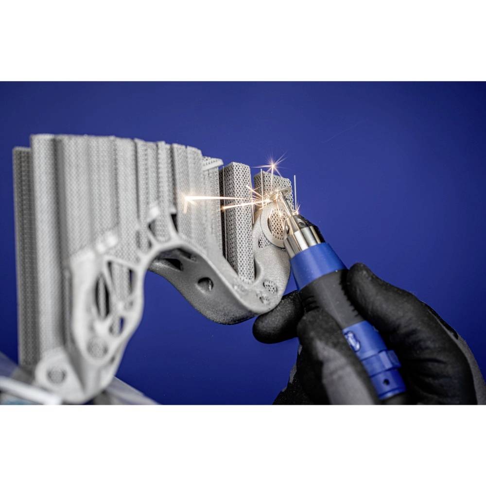 Une personne travaille sur une pièce métallique complexe imprimée en 3D à l'aide d'un outil bleu, des lumières scintillantes signalent un travail de précision.