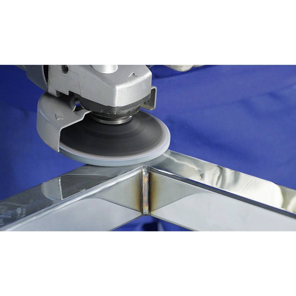 Une meuleuse traite un angle soudé d'un bâti métallique. Un tissu bleu est visible en arrière-plan.