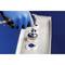 Une personne portant des gants bleus utilise une machine pour poinçonner des trous métalliques dans un récipient rectangulaire.