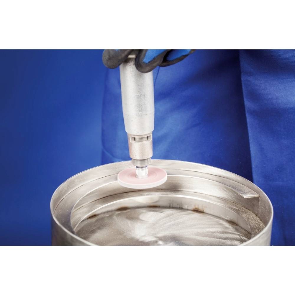 Une personne vêtue de vêtements de travail bleus polit la surface d'un fût métallique à l'aide d'un outil électrique.