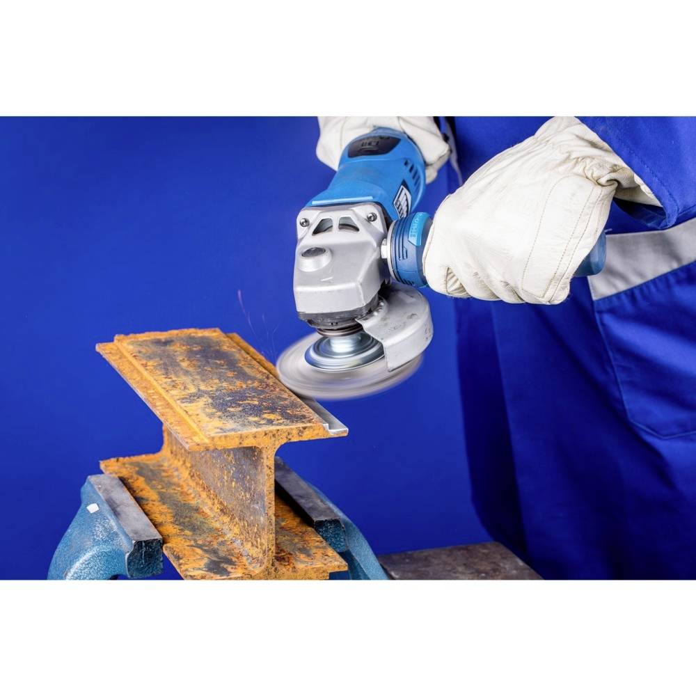Une personne enlève de la rouille d'un support métallique à l'aide d'une meuleuse d'angle. Elle porte des gants de travail et des vêtements de protection bleus.