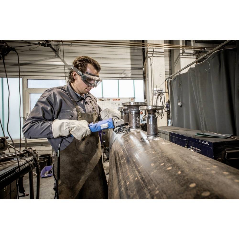 Un ouvrier en tenue de protection meule un tube métallique dans un atelier. Des outils et des machines sont visibles en arrière-plan.