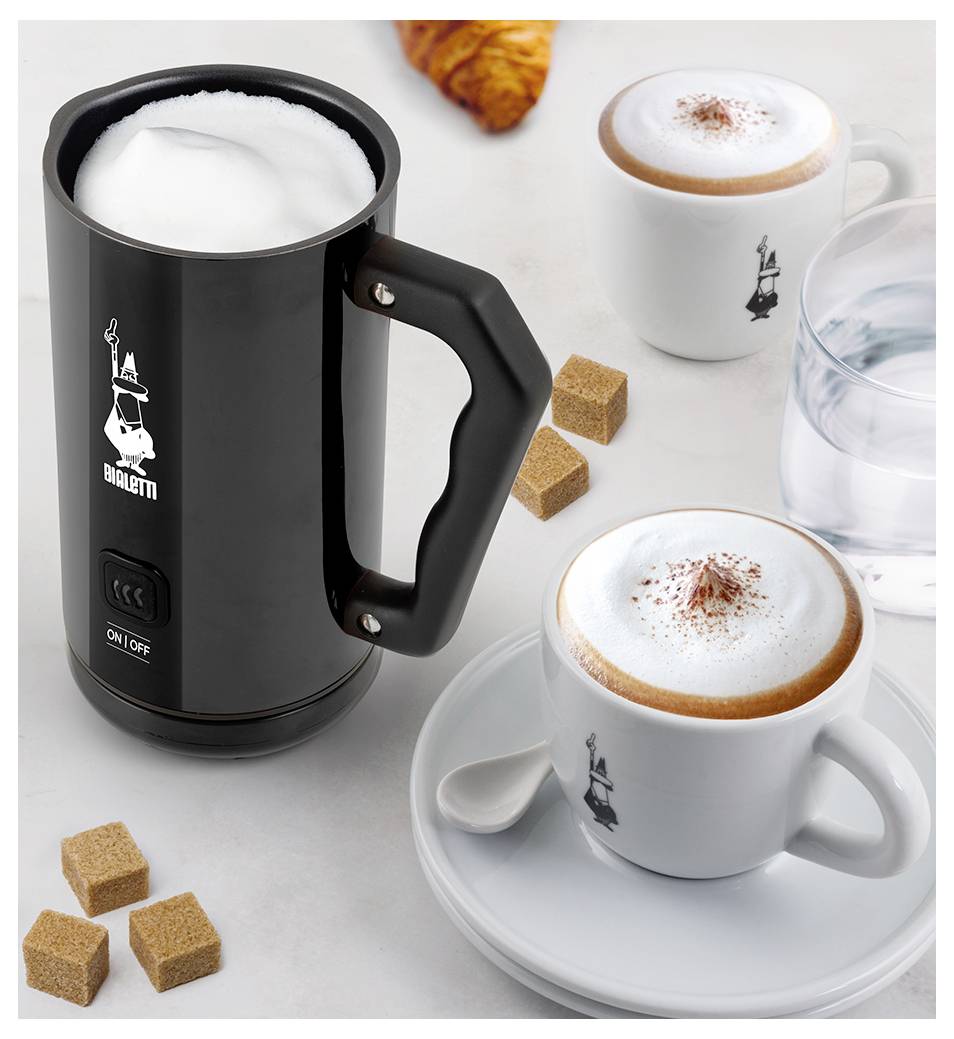Un mousseur à lait Bialetti à côté de deux tasses de cappuccino, des morceaux de sucre et un croissant sur une table.
