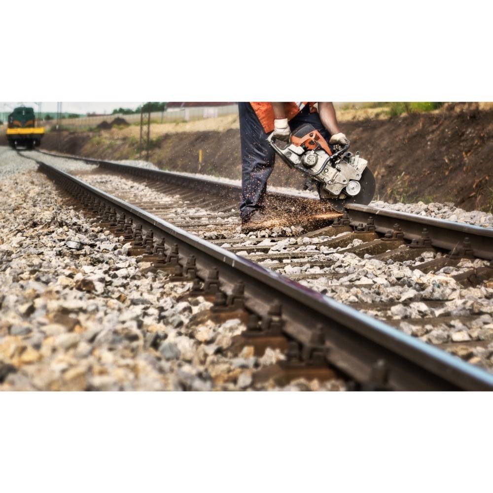 Une personne découpe des rails avec une tronçonneuse sur une voie ferrée. Un train s'approche en arrière-plan. Scène de chantier.