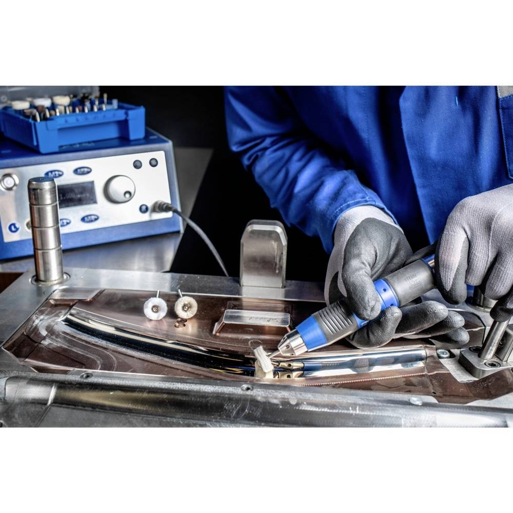 Une personne vêtue de bleu effectue un travail de soudure précis sur des pièces métalliques, avec un dispositif technique en arrière-plan.