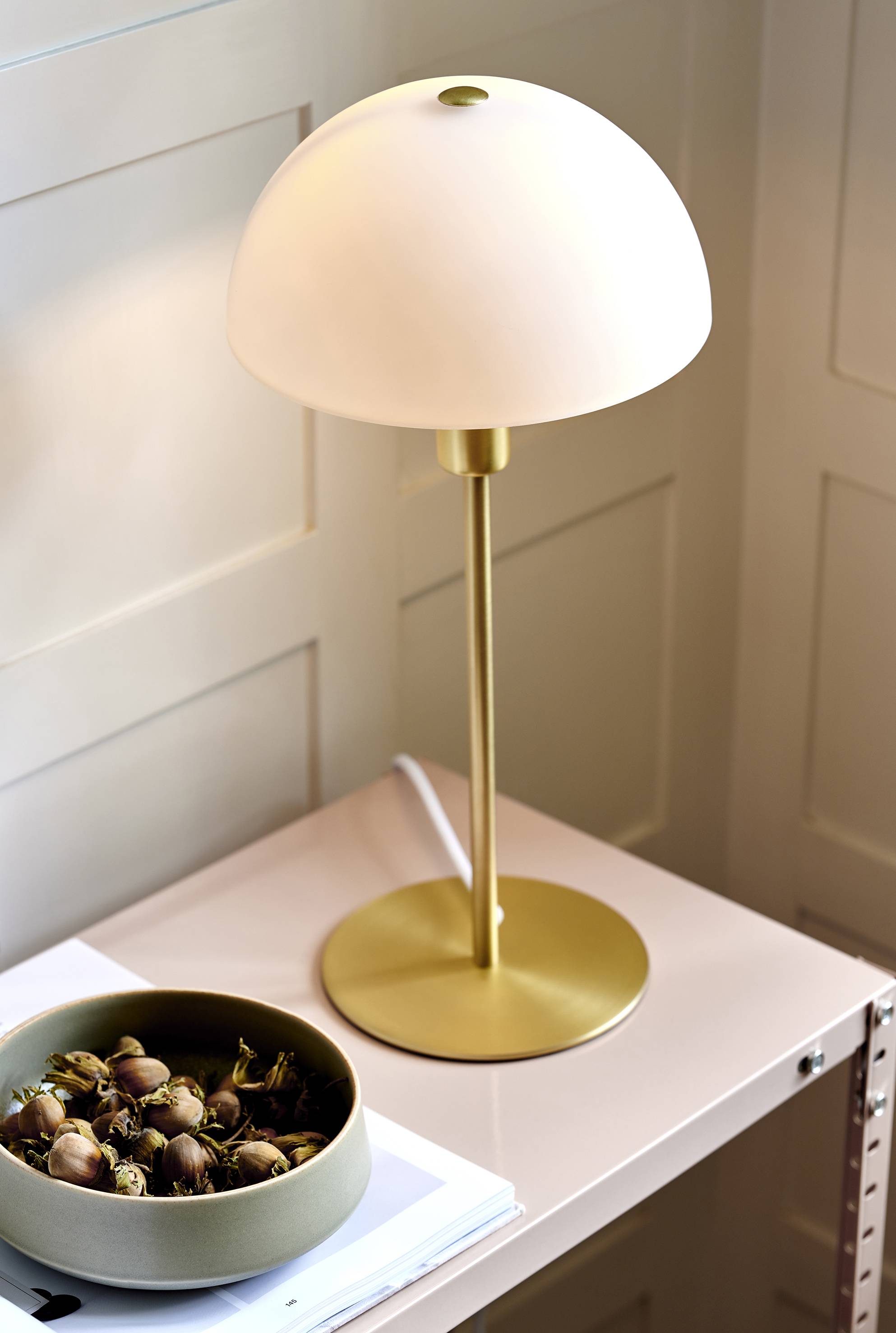Une lampe de table dorée à abat-jour rond est posée sur une table à côté d'un bol de noisettes. L'environnement paraît minimaliste.