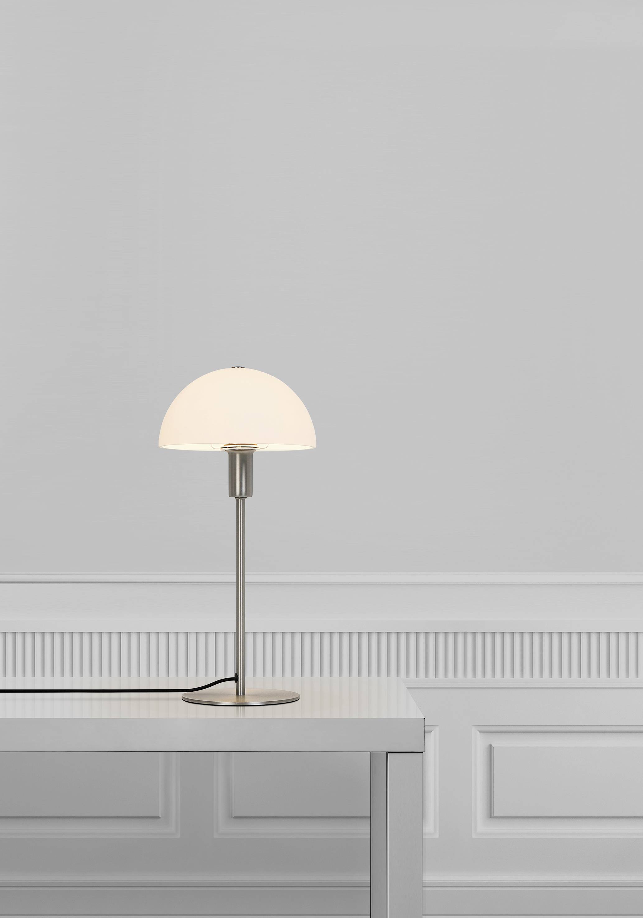 Une lampe de table moderne avec un abat-jour blanc est posée sur une table blanche simple devant un mur gris clair orné de panneaux décoratifs.