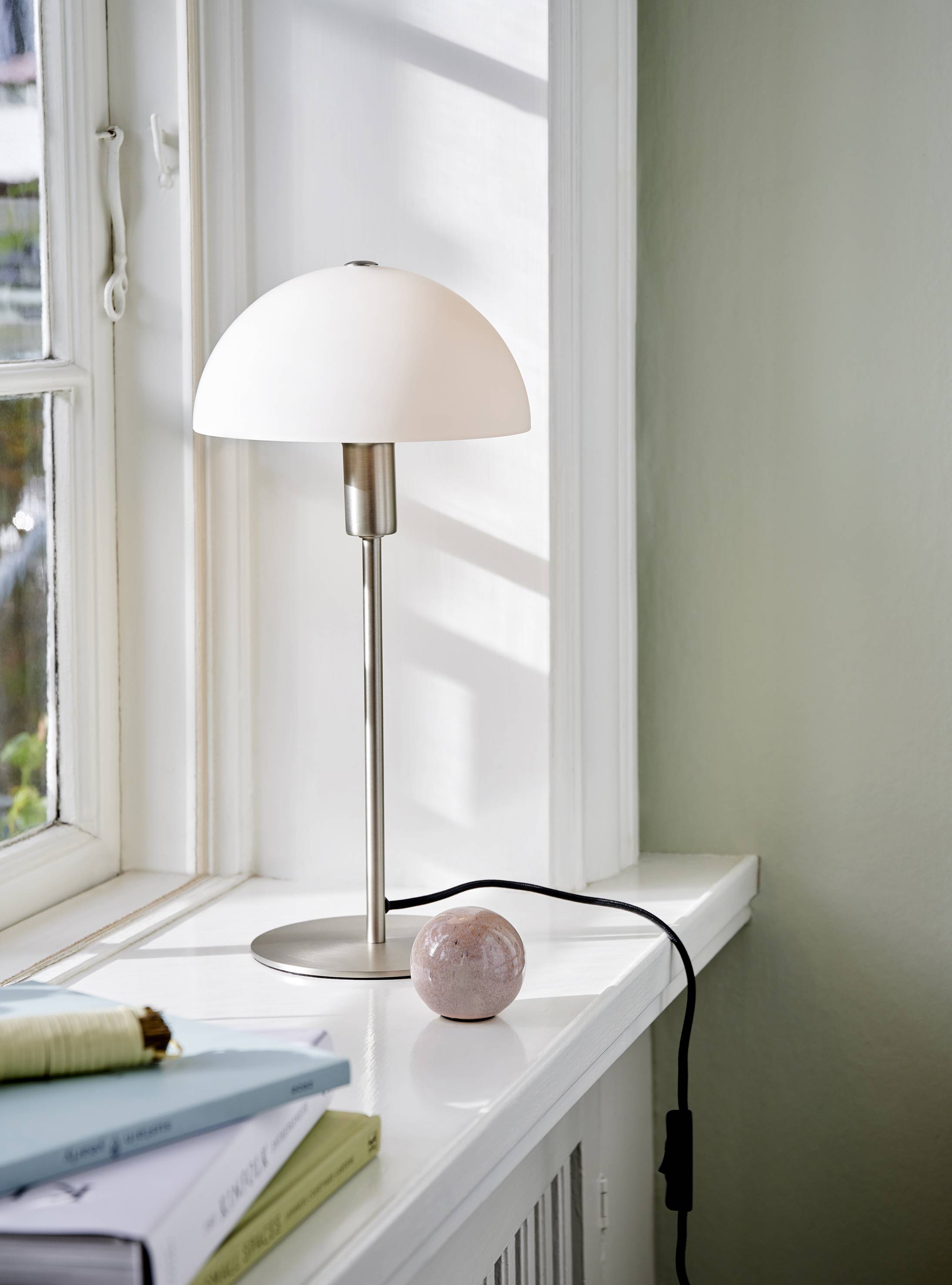 Une lampe de table moderne avec un abat-jour blanc est posée sur un rebord de fenêtre. À côté de la lampe se trouve un objet rond rose et des livres.