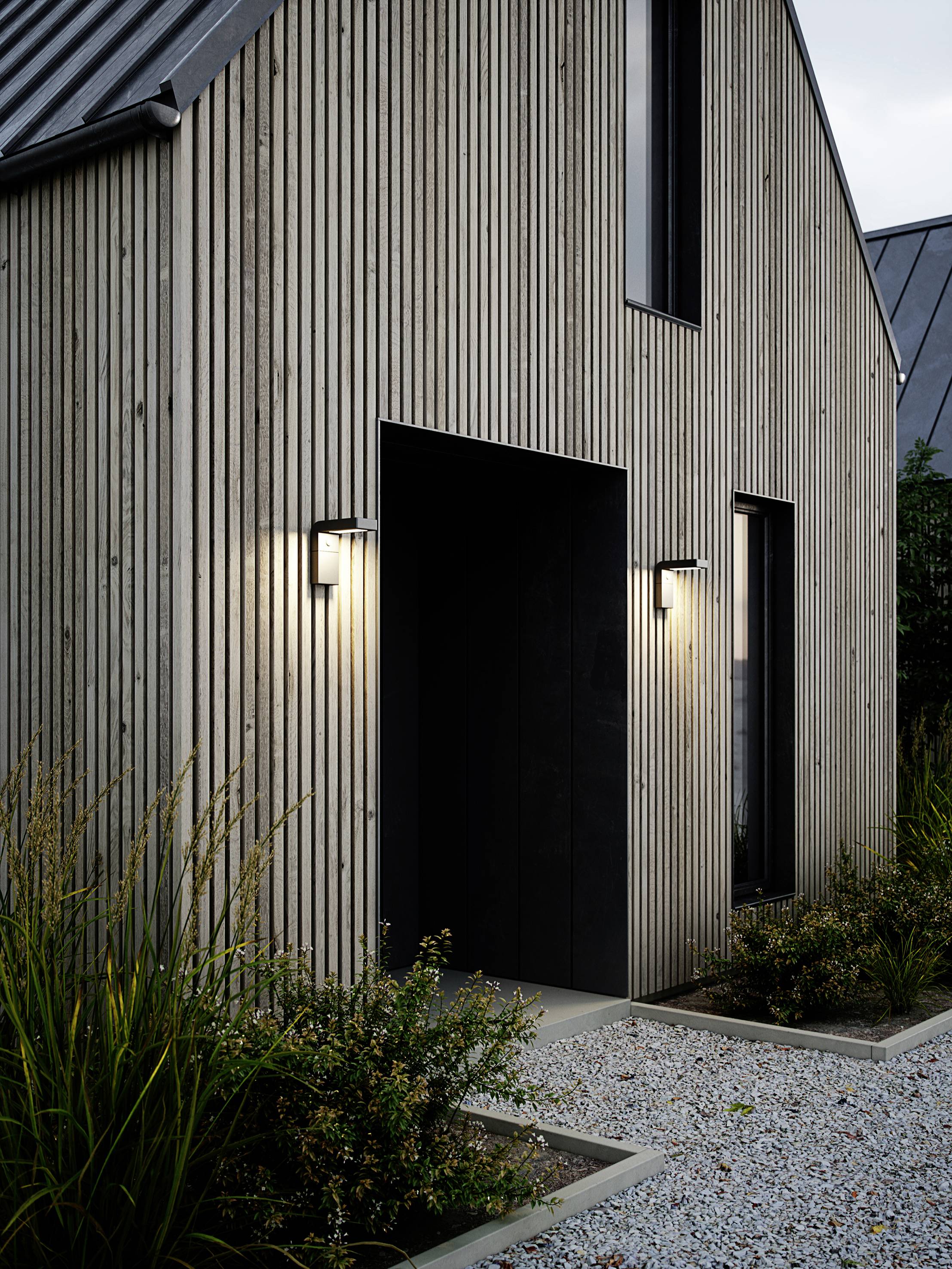 Une maison moderne avec un revêtement en bois, éclairée par des lampes extérieures. Un chemin de gravier mène à l'entrée, entouré de plantes.