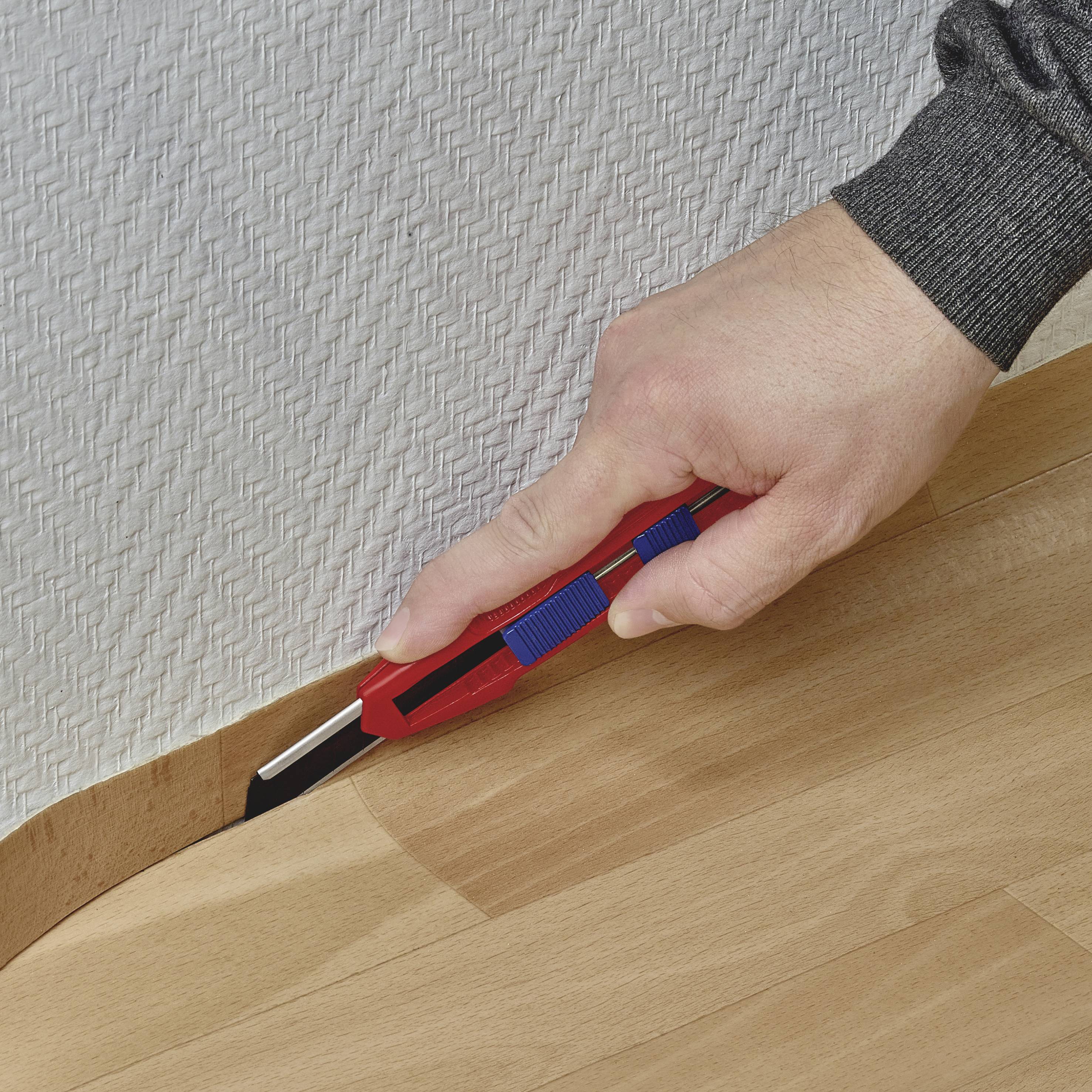Une main coupe un morceau de tapis avec un cutter le long d'un revêtement de sol.