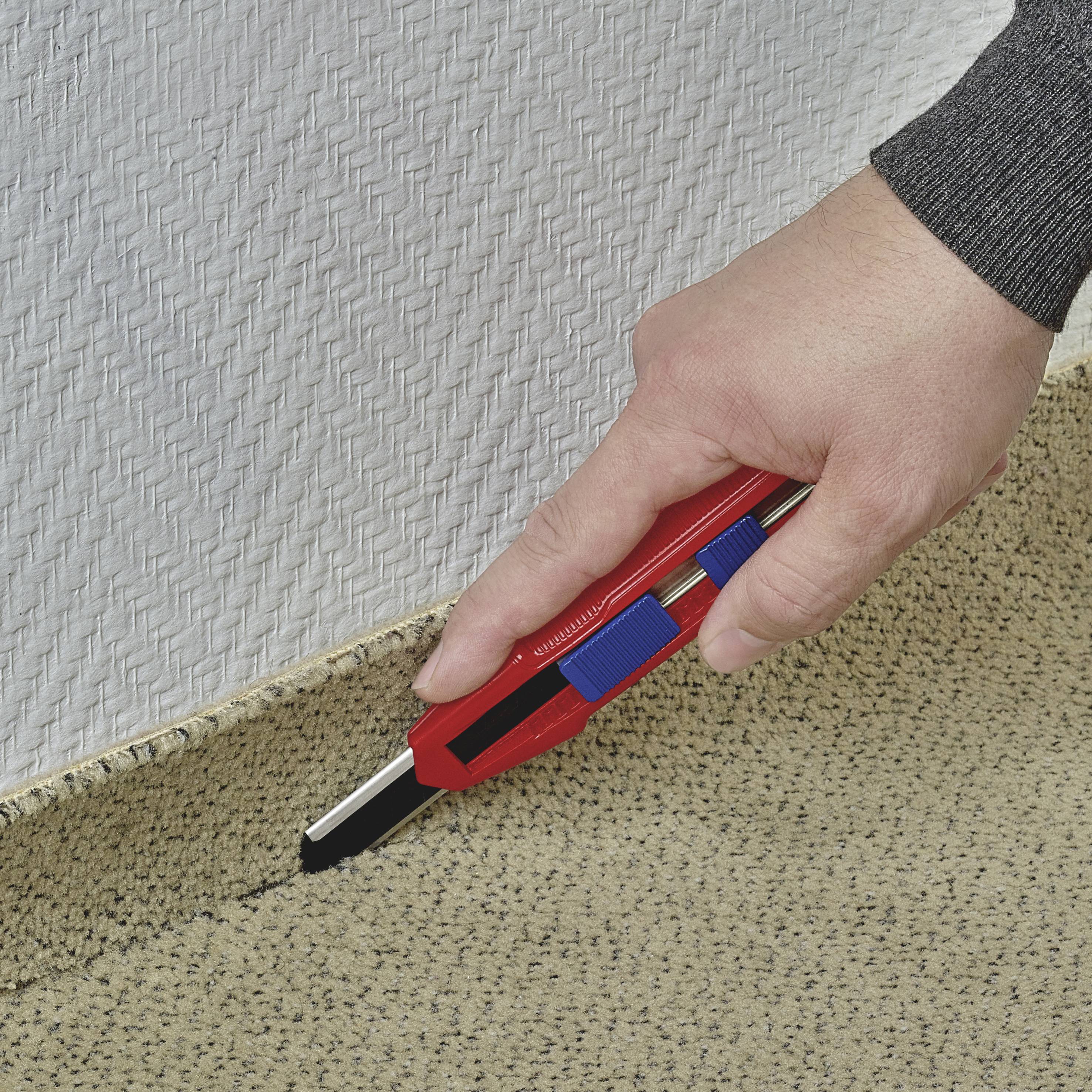 Une main coupe un revêtement de sol en moquette le long d'un bord à l'aide d'un couteau à tapis. La moquette présente une texture beige.