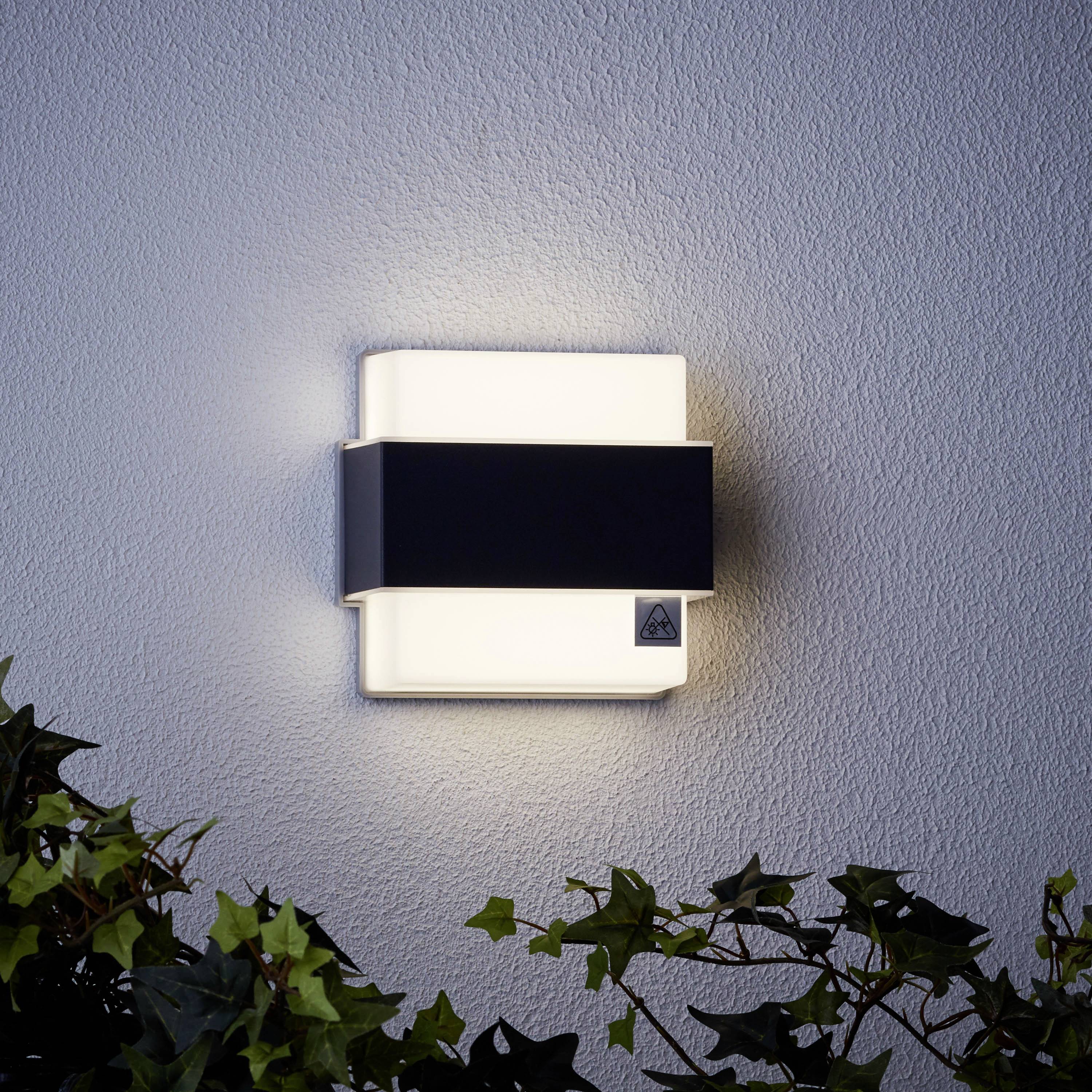 Un luminaire mural moderne au design géométrique éclaire un mur gris. Des plantes vertes sont visibles en bas.